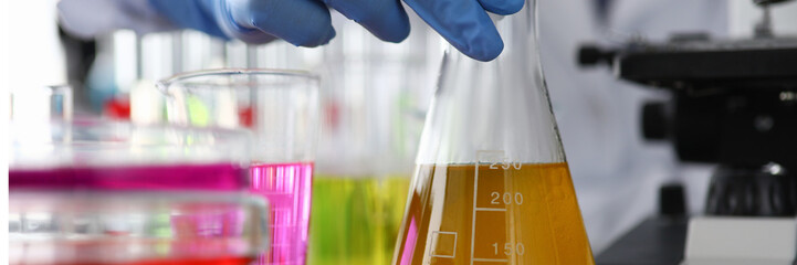 Close-up of chemists hand holding beaker with yellow liquid for further expertise. Lots of lab working stuff on table. Worker wearing solid protection. Medical analysis concept