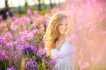 Young beautiful blonde girl gathers pink flowers in the spring blooming field at sunset.