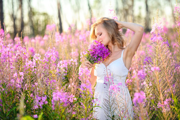 Young beautiful blonde girl gathers pink flowers in the spring blooming field at sunset.