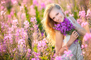 Young beautiful blonde girl gathers pink flowers in the spring blooming field at sunset.