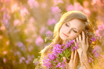 Fototapeta premium Young beautiful blonde girl gathers pink flowers in the spring blooming field at sunset.