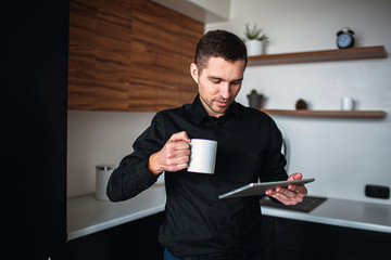 Young businessman work in kitchen. Entrepreneur stand alone, hold tablet and look at it. Businessman has productive day. Coffee break or rest.