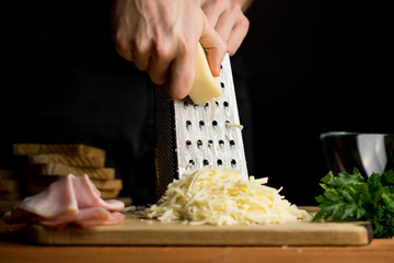 A person grinding cheese on an aluminum grater preparing cheese for sandwiches with ham and herbs