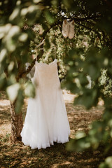 White wedding dress and unusual wedding sneakers hanging on tree in country garden full of morning sunlight. Focused on wedding dress,the surrounding area is slightly out of focus with leafy bokeh.