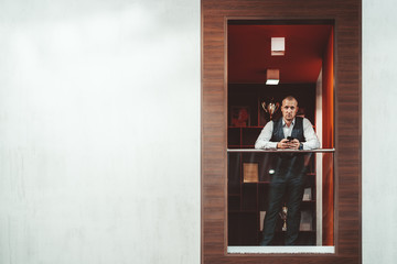 Serious handsome hispanic man entrepreneur is leaning against balcony railing while using his smartphone in a recess of a modern office chillout area, with a copy space place on the left for a message
