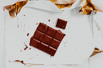 large chocolate bar broken into squares on dirty white paper with chocolate chips and gold foil around the edges.