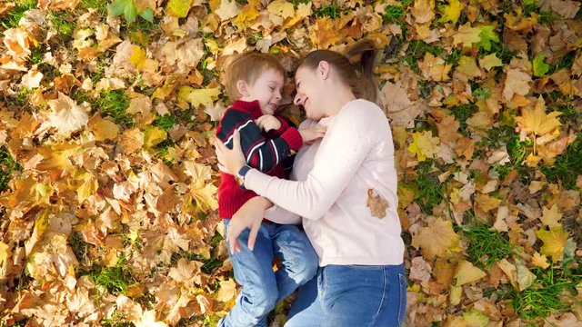 4k video of little toddler boy with mother playing, tickling each other, hugging and kissing while lying on yellow leaves and grass in autumn park