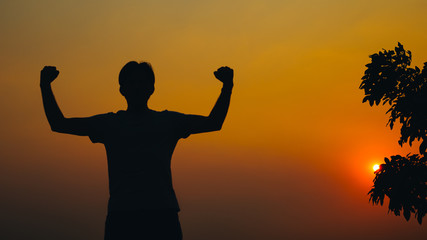 Silhouette young man with hands raised standing high up during sunset and tree background