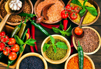 Wooden bowls of various lentils with red and green hot chilli peppers with leaves and tomatoes on a wooden background. Top view.