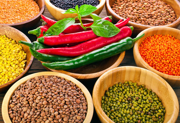 Wooden bowls of various lentils with red and green hot chilli peppers with leaves and tomatoes on a wooden background. Top view.