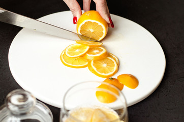 Food black background. On a white stone board in the hands of a girl, a knife cuts a lemon. Slices of lemon are piled in a glass jar and sprinkled with sugar. In the foreground is a whole lemon.