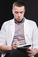 Portrait of a young intern doctor. A man sits on a chair with a notebook and pen in his hands and writes something. Making a diagnosis. Studio portrait