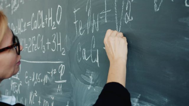 Close-up of woman's hand writing formulas on blackboard in college classroom teaching chemistry. Modern education, people and knowledge concept.