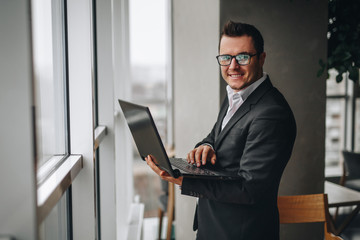 rich guy holds a laptop in his hands and works, standing by the window