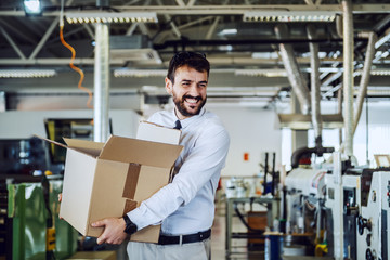 Smiling caucasian bearded graphic engineer in shirt and tie walking in printing shop and relocating box. In background are printing machines.