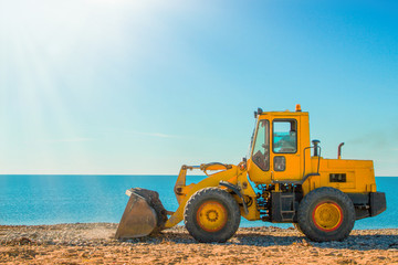 Obraz na płótnie Canvas Yellow bulldozer on the background of clear blue sky and seashore. Copy space and light from the left side