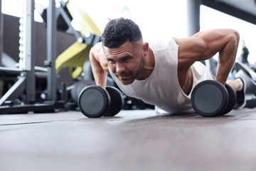 Fit and muscular man doing horizontal push-ups with dumbbells in gym.