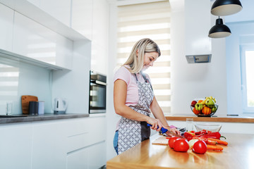 Smiling attractive worthy Caucasian blonde woman in apron cutting vegetables while standing in kitchen. On kitchen counter are carrots, tomatoes and peppers.