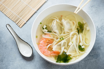 Bowl of panasian noodle soup with wontons and shrimps, high angle view on a light-blue stone background, studio shot