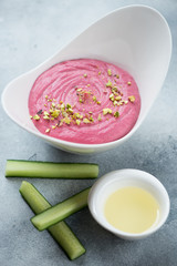 White bowl of hummus with addition of beetroot and cucumber slices, vertical shot on a light-blue stone surface