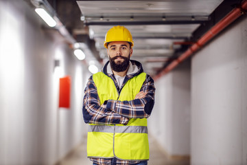 Cute Caucasian bearded construction worker with safety helmet on head in vest standing with arms...