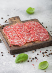 Classic salami slices with basil and pepper on wooden chopping board and light kitchen table background.
