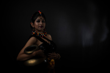 Obraz na płótnie Canvas Fashion portrait of Indian Bengali brunette woman in Indian tribal dress and handmade ornaments holding metal pot in dark copy space studio background. Indian lifestyle and fashion photography.