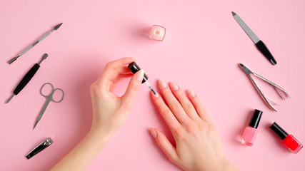 Woman making manicure herself. Female hands with nail polish and manicure tools on pink background, view from above. Self-care beauty treatment concept