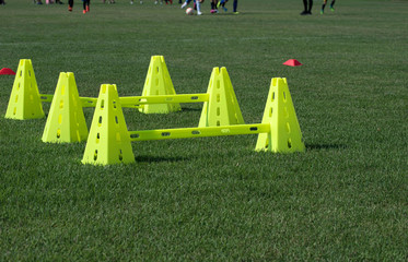 High & low plastic lime green pyramid  cones with bars attached placed on a football pitch ready for an afternoon practice in a school soccer field. Youth players in action in a background. - Powered by Adobe
