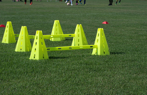 High & low plastic lime green pyramid cones with bars attached placed on a football pitch ready for an afternoon practice in a school soccer field. Youth players in action in a background.