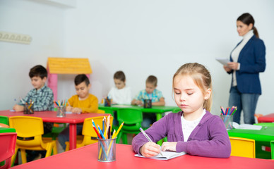 Portrait of little assiduous girl with pen and notebook at lesson in elementary school