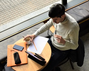 Top view of a young man putting on his glasses to work with a tablet while enjoying a cup of coffee in front of a window
