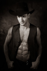 Portrait of handsome cowboy looking at camera wearing hat with open waistcoat revealing defined...
