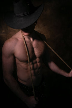 Shirtless cowboy wearing hat covering his face, holding rope around his neck with defined pecs and muscular sixpack abs