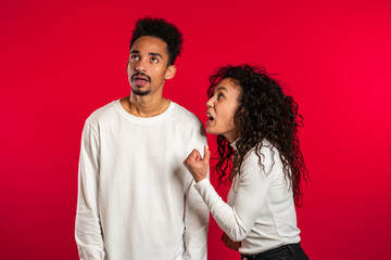 Young african woman emotionally screaming at her husband or boyfriend on red background in studio. Bored man rolling his eyes. Concept of conflict, problems in relationships.