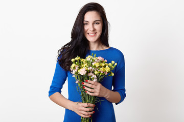 Studio shot of beautiful brunette girl holding bouquet of flowers, attractive woman with big bunch of flowers, dark haired female dressed blue dress, posing over white background, expressing happyness