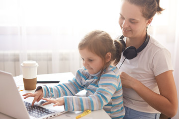 Close up portrait of woman and young cute girl sitting in front of opened laptop, mother holding her daughter on knees, female child dresses stripped shirt playing computer game with her mommy.