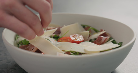 man hand adding parmesan flakes to make salad with arugula, tomatoes and prosciutto