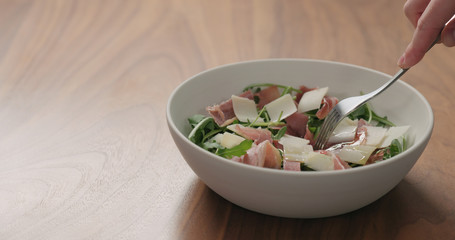 man hand pick salad with arugula, prosciutto and cherry tomatoes in white bowl on walnut table