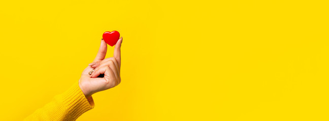Hand holding small red heart over yellow background, panoramic mock-up