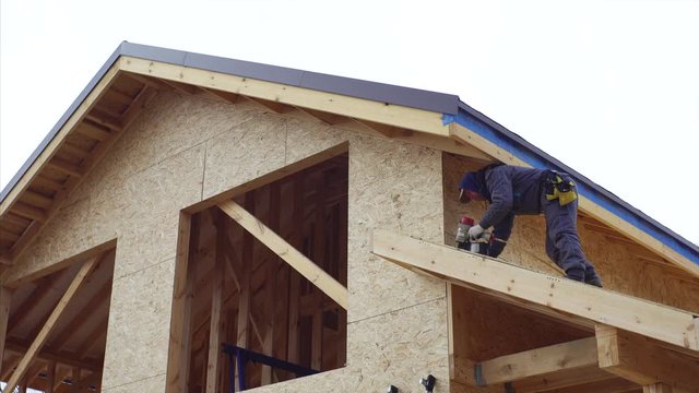 View from below of builder using hydraulic hammer while covering trussing of small roof with plywoods. Residential frame house under construction