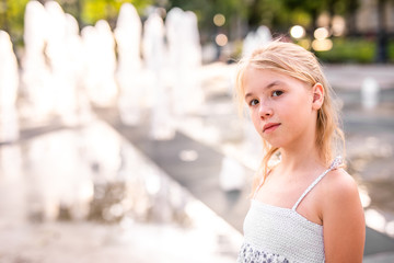 Blonde happy child girl in long purple dress running between water flow in city summer park. Child activity concept