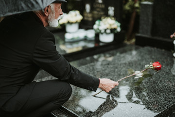 Elegant sad elderly man standing on the rain with umbrella and grieves at the grave of a loved...