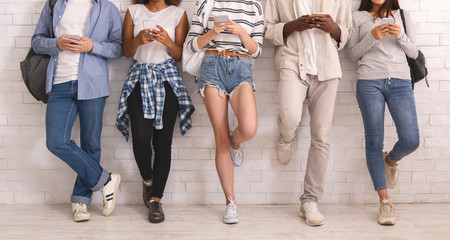 Cropped of multiethnic friends uisng smartphones, leaning on white wall
