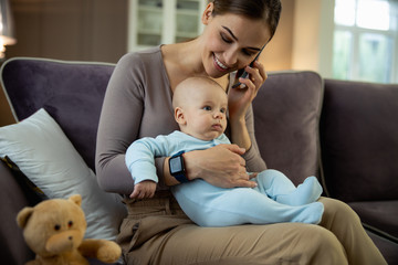 Smiling pretty woman sitting with baby and using smartphone