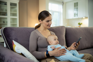 Smiling young mother sitting with kid and using mobile phone