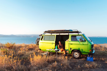 Young attractive female sitting in old timer camper van on a hill above the beach looking at the...