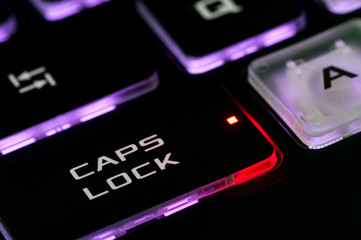 The Caps lock key on a backlit keyboard. The red illuminated dot on the key indicates that the capslock is turned on. Closeup, selective focus.