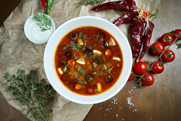 Top view on tasty Solyanka thick, spicy and sour Russian soup with olives, lemon and sausages in white bowl on wooden background.