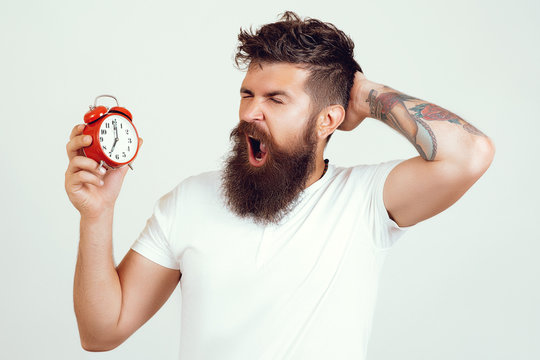 Handsome man yawning holds vintage alarm clock over isolated background. Hipster with tattoo gets up with alarm clock. Hipster man with alarm clock. I should go to bed earlier.